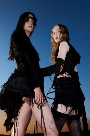 Two models in avant-garde black couture garments posing outdoors against a dusk sky from a low angle.