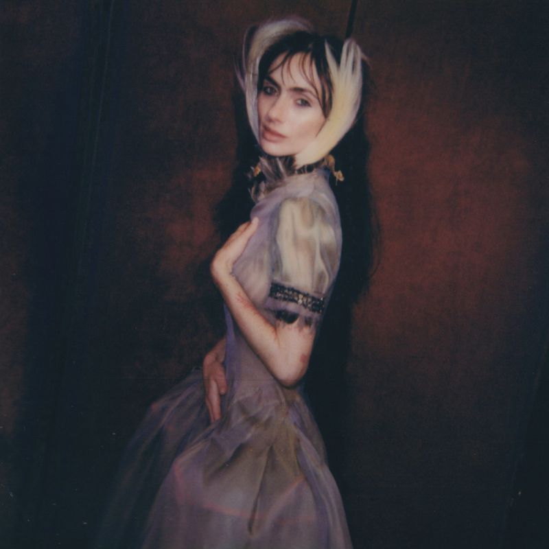 Woman in a sheer, vintage-style dress with feather headpiece poses against a dark red background in a medium shot.