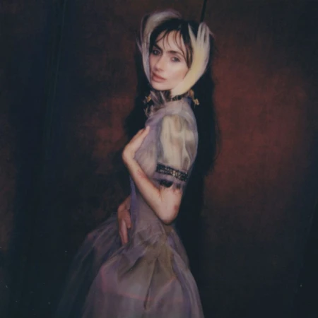 Woman in a sheer, vintage-style dress with feather headpiece poses against a dark red background in a medium shot.