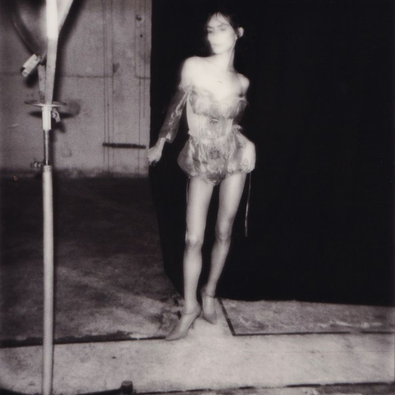 Woman in a corset and heels stands in front of a dark background in a blurry black-and-white studio fashion photograph.