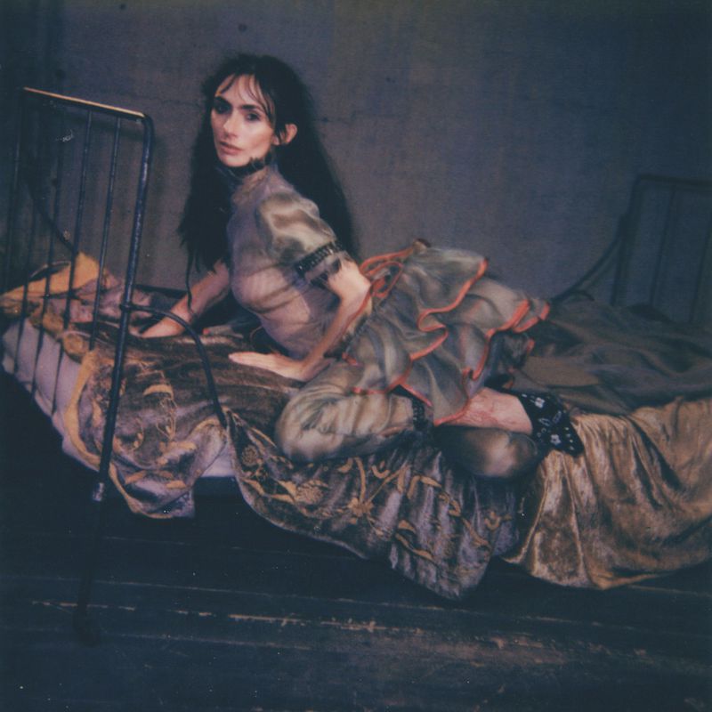 A woman with long dark hair, dressed in a grey sheer dress with a ruffled skirt, poses on a bed with metal frame against a grey wall.