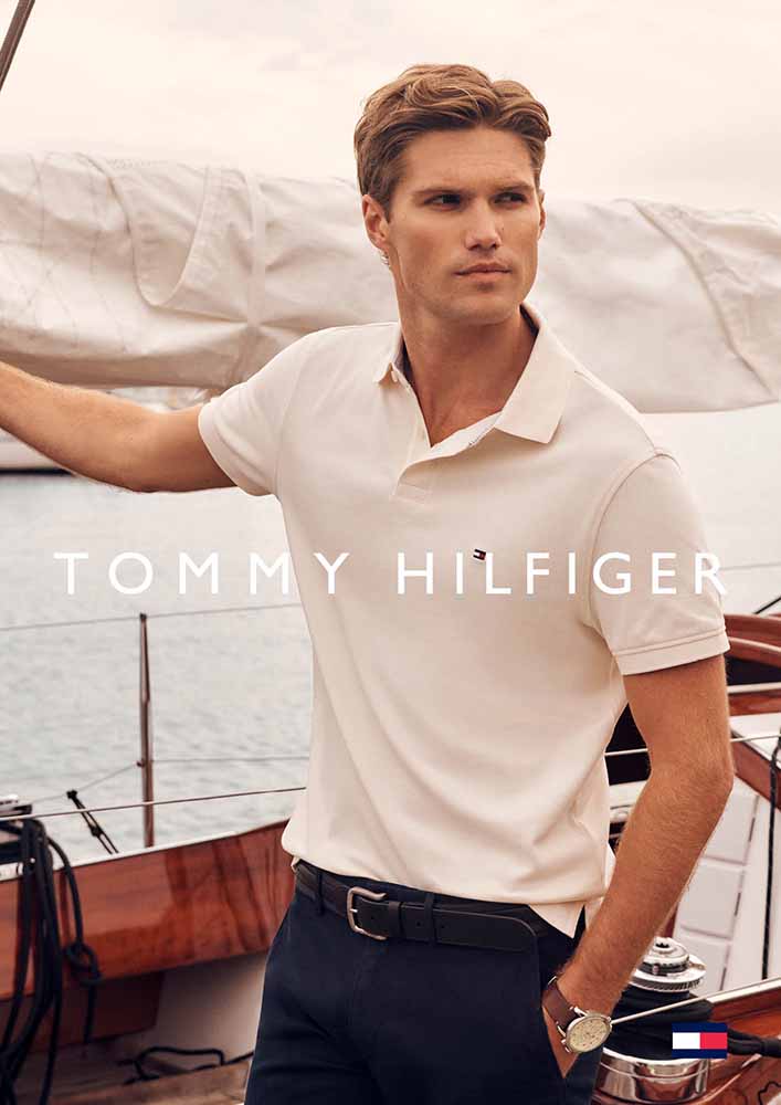 Handsome blond male model poses wearing a white polo shirt in a sailing scene, with branding visible.