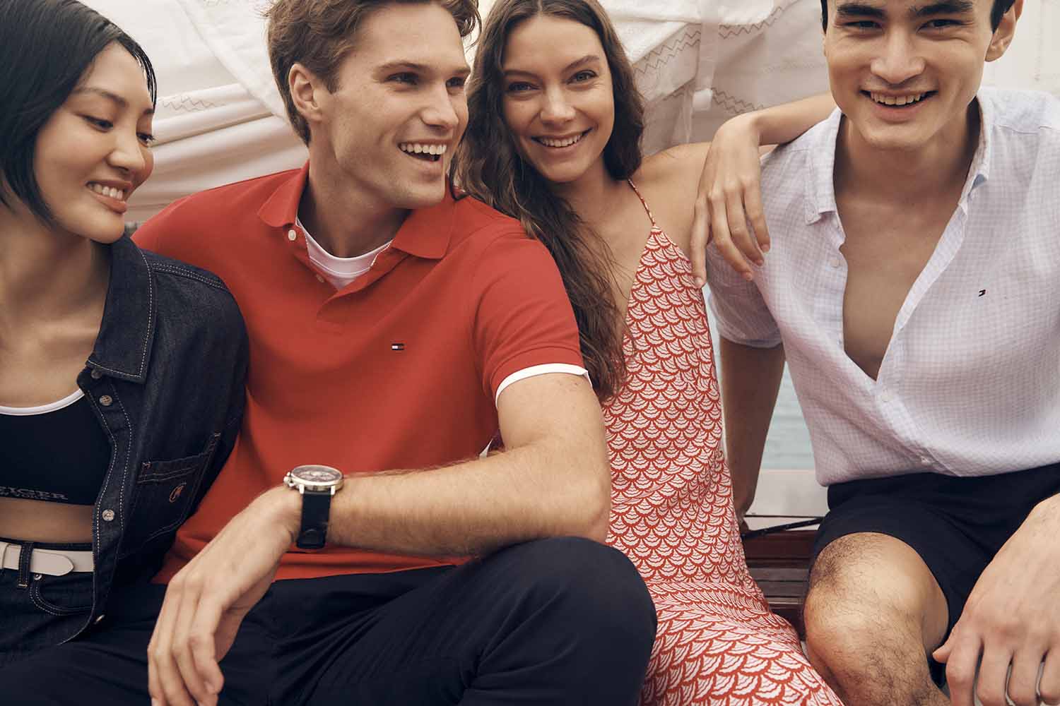 Four young adult friends sit together smiling, wearing casual clothing: polo shirt, dress, button-down shirt, and jacket, in an indoor setting.