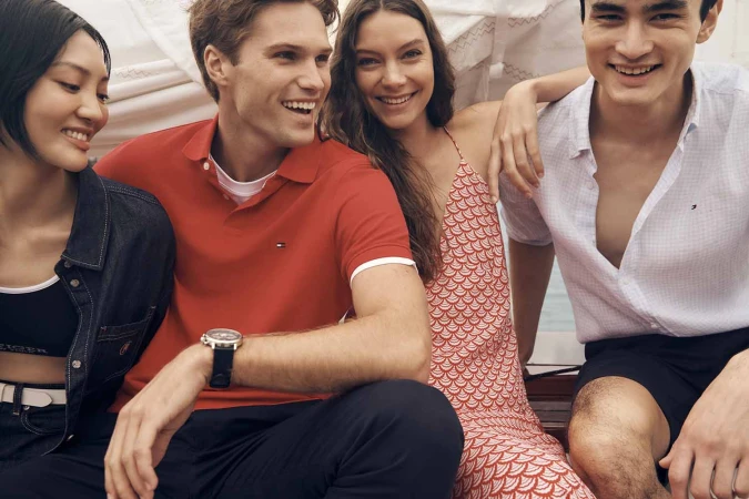 Four young adult friends sit together smiling, wearing casual clothing: polo shirt, dress, button-down shirt, and jacket, in an indoor setting.