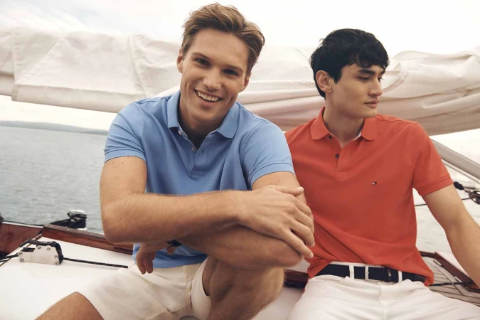 Two young men are on a sailboat. One is smiling wearing a blue polo shirt, while the other, in an orange polo shirt, looks away.