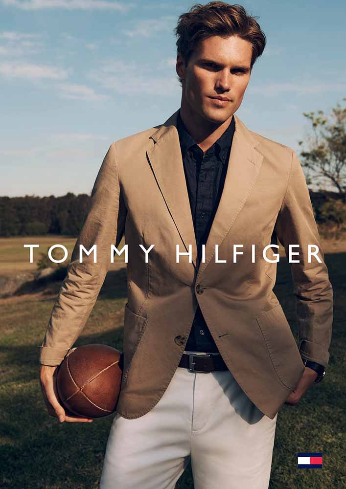A male model in a tan blazer and white pants holds a football on a grassy field with Tommy Hilfiger logo and lettering at center.