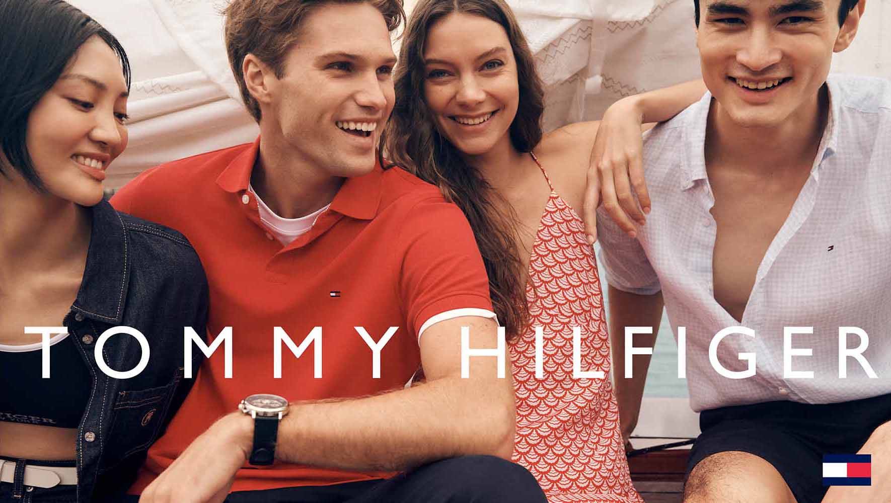 Four young adults of diverse ethnicities smile together outdoors on a bright day, wearing casual summer clothes, promoting the Tommy Hilfiger brand.
