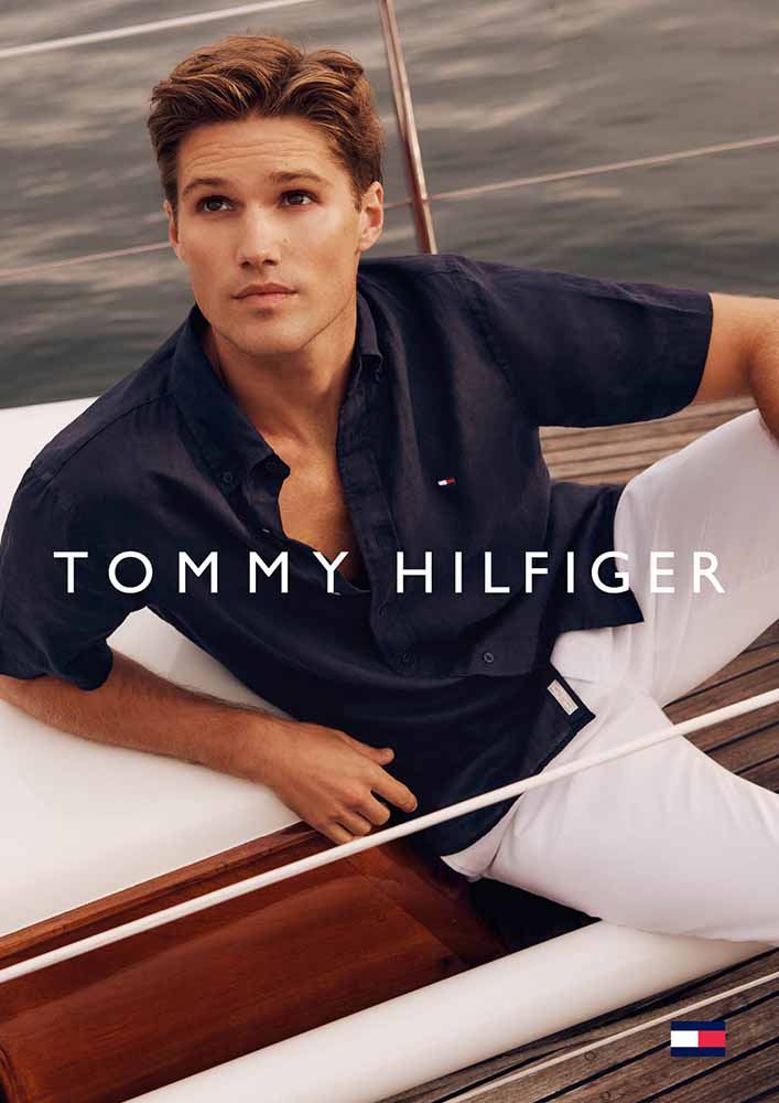Man in unbuttoned navy blue shirt and white pants poses on yacht. Tommy Hilfiger logo. Fashion and Lifestyle photography.