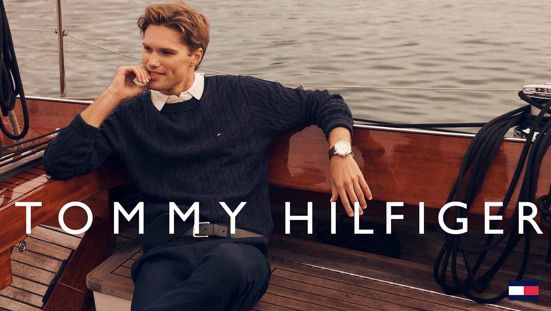 A blond male model relaxes on a wooden boat wearing a Tommy Hilfiger sweater, watch, and belt, with the brand's logo on the sweater.
