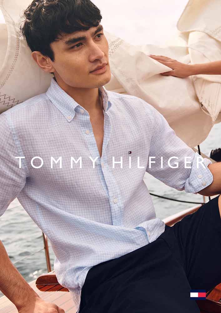 An Asian man wearing a light-blue checkered shirt poses on a boat, looking off-camera with a relaxed expression in soft, natural lighting.
