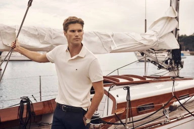 Male model in a white polo shirt and dark trousers standing on the deck of a sailboat for Tommy Hilfiger.