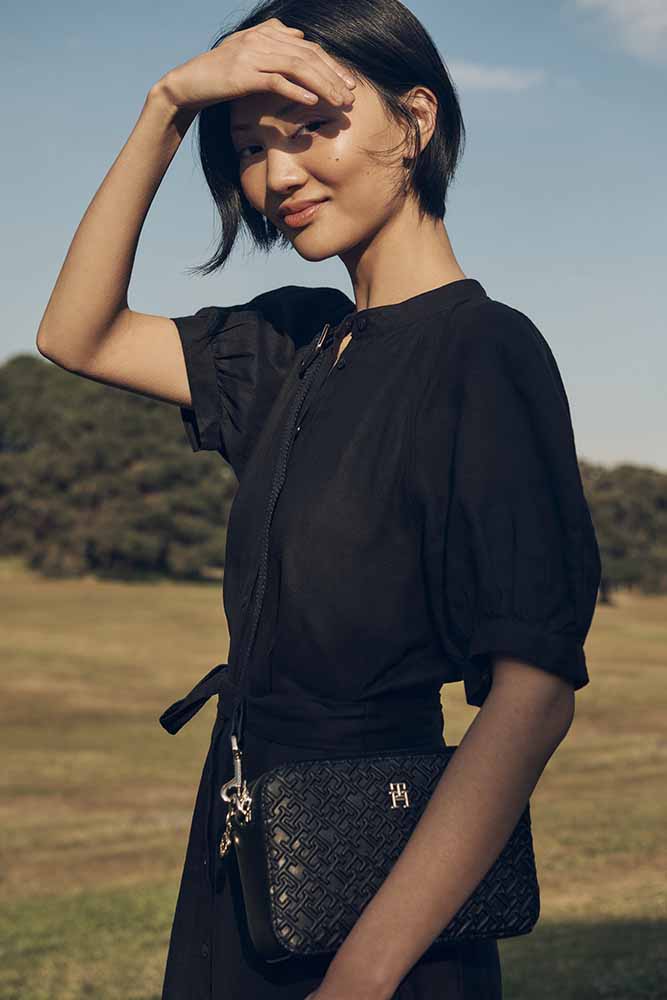Asian woman in black dress shields eyes in outdoor fashion shot with textured black crossbody bag, soft focus green background.