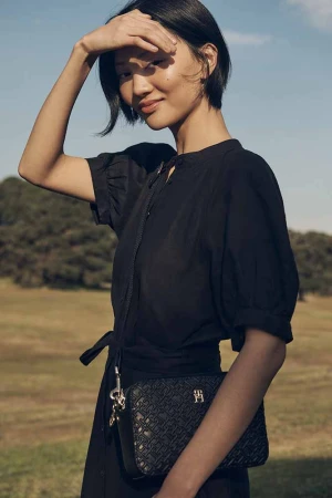 Asian woman in black dress shields eyes in outdoor fashion shot with textured black crossbody bag, soft focus green background.