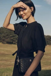 Woman in black dress with a black crossbody bag shielding her eyes from the sun outdoors