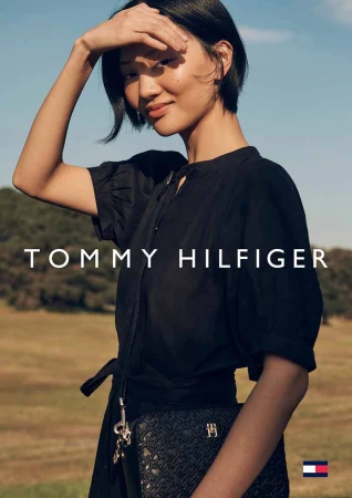 Model with dark hair wearing a black dress and holding a crossbody bag outdoors against a grassy landscape