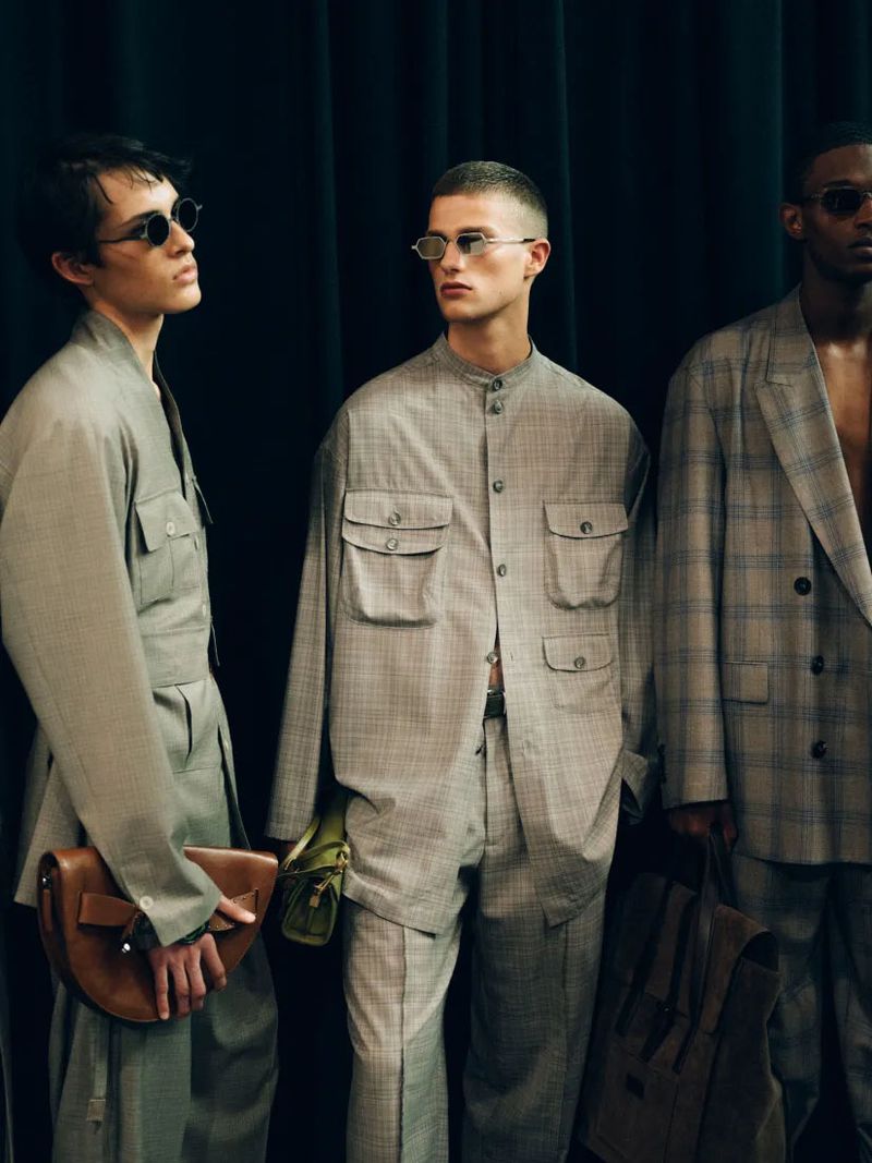 Three male fashion models pose indoors against a dark curtain backdrop, wearing matching grey suits and sunglasses, holding purses.