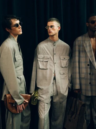 Three male fashion models pose indoors against a dark curtain backdrop, wearing matching grey suits and sunglasses, holding purses.