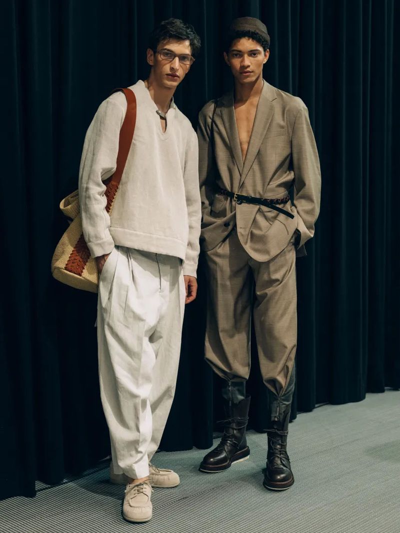 Two stylish men pose in relaxed tailored outfits, one in a sweater with shoulder bag, the other in a suit with vintage riding boots.