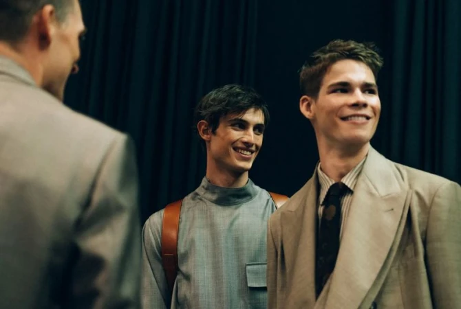 Three men in suits are smiling in front of a dark background, dressed in blazers and a turtleneck shirt, in a fashion forward aesthetic.