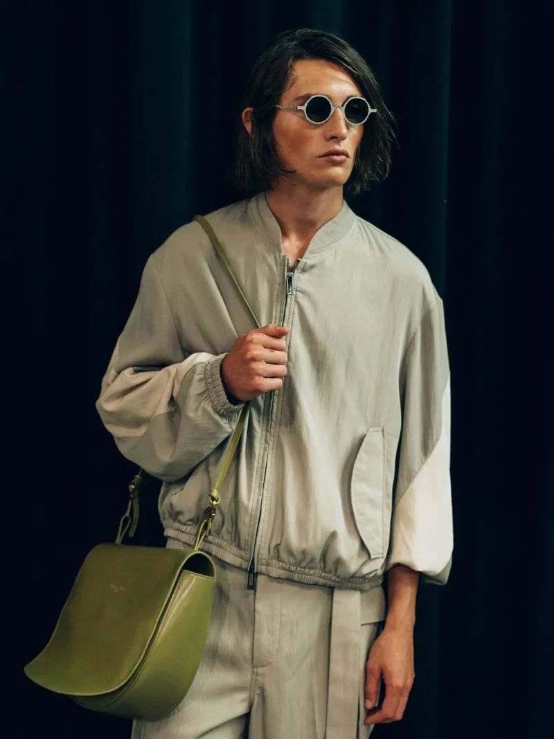 An adult male model with medium-length hair wearing sunglasses, a light grey bomber jacket, and striped trousers with an olive bag on a black background.