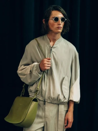 An adult male model with medium-length hair wearing sunglasses, a light grey bomber jacket, and striped trousers with an olive bag on a black background.