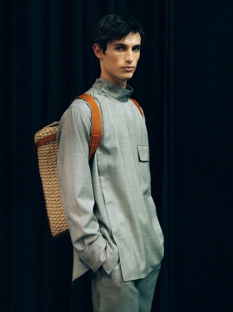 A young adult man in a grey suit and woven backpack poses indoors against a dark curtain backdrop in a fashion editorial photograph.