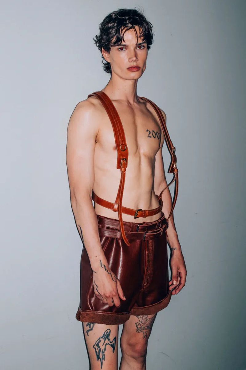 A lean adult male model in leather shorts and suspenders strikes a pose in a studio environment with tattoos and dramatic lighting.