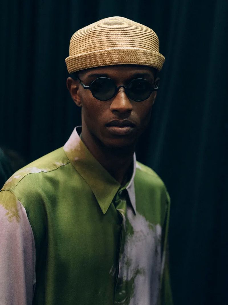 Close-up of a dark-skinned adult male wearing a straw hat and sunglasses, posing against a dark green curtain backdrop indoors.