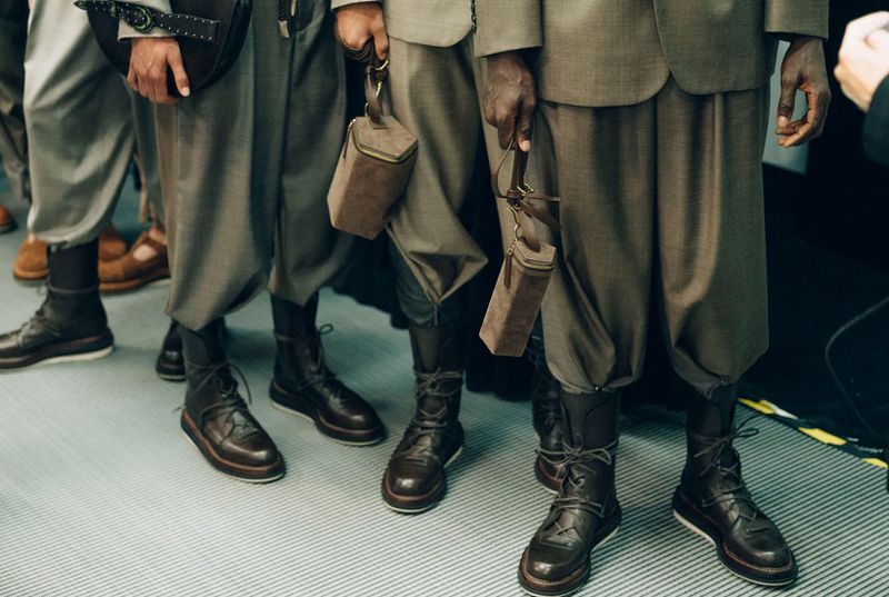 Fashion show with models in tailored suits and lace-up boots, holding small pouches, captured indoors under diffused lighting.