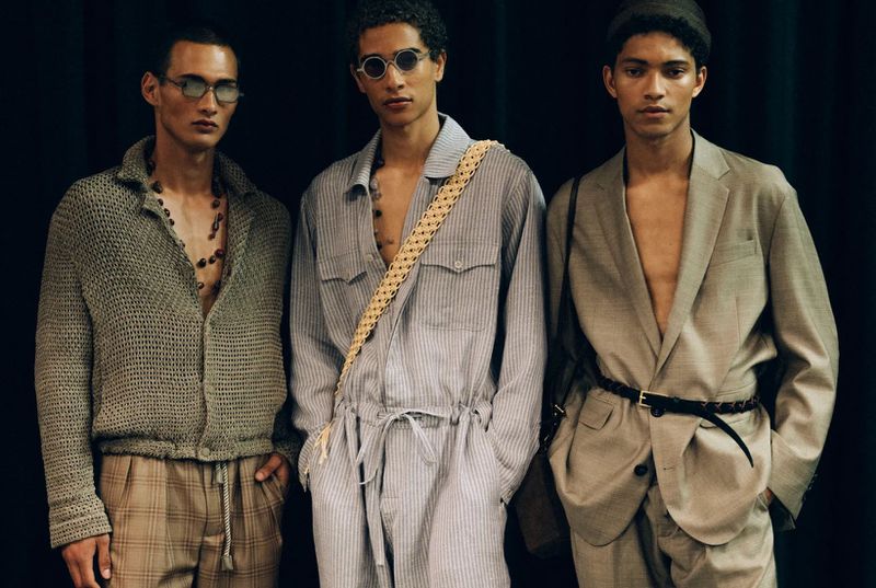 Three fashionably dressed adult male models pose against a dark backdrop, showcasing contemporary menswear and accessories in a studio setting.