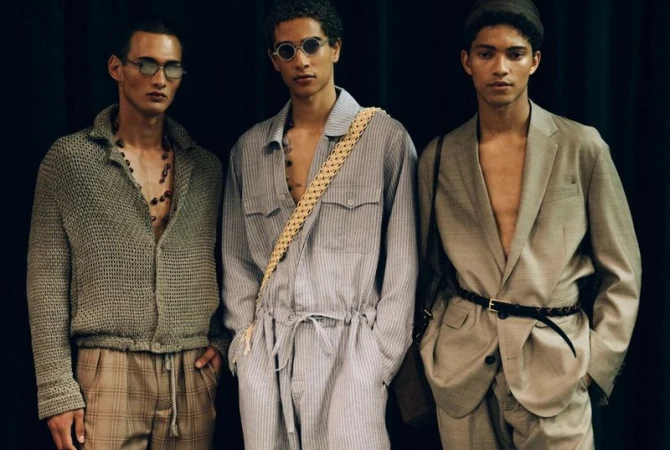 Three fashionably dressed adult male models pose against a dark backdrop, showcasing contemporary menswear and accessories in a studio setting.