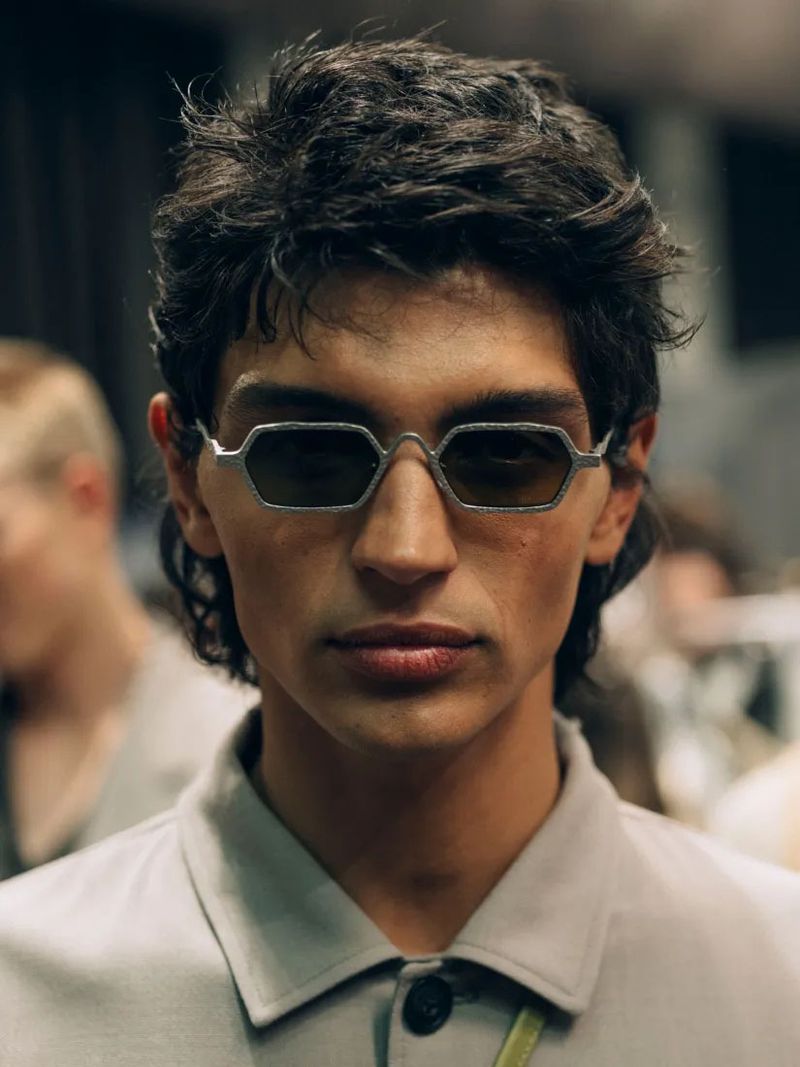 Portrait of a young adult male wearing geometric sunglasses, styled hair, and a neutral button-up shirt in a backstage environment.