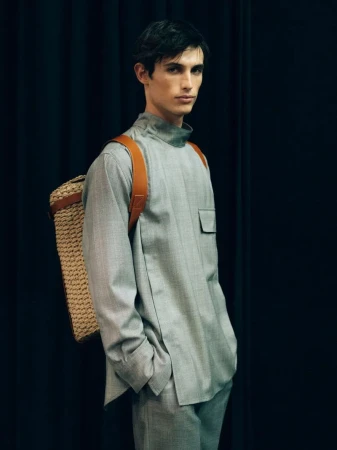 Male model wearing grey tunic and trousers carrying a woven backpack in front of black drapes.