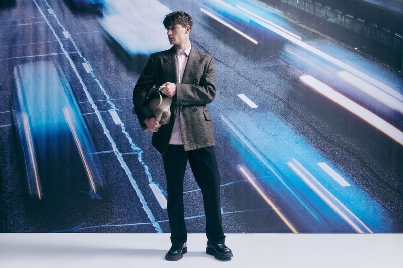 Man in suit jacket holds bag, posing against a backdrop of blurred city lights, in a studio setting with neutral lighting.