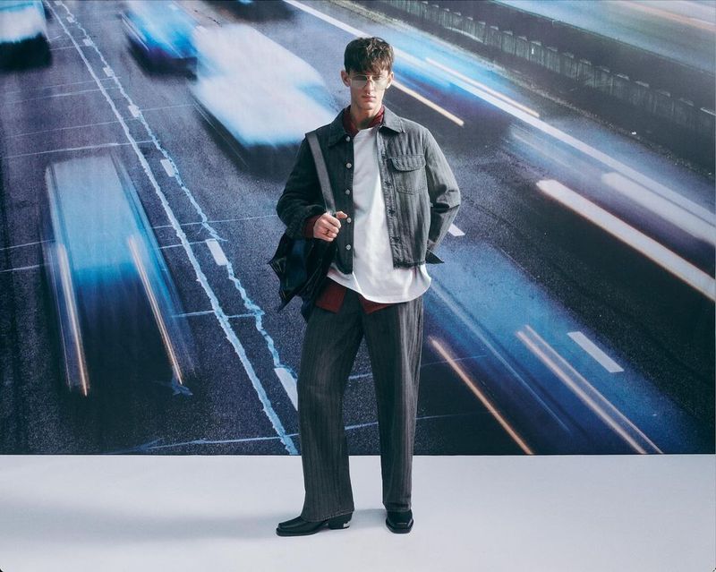 Man in denim jacket and grey trousers poses against blurred urban road backdrop. Fashion shot with male model wearing casual clothing.