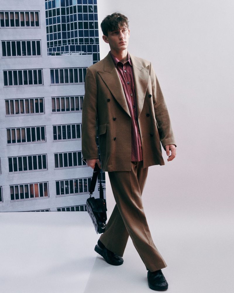 A young male fashion model in a tan double-breasted suit walks confidently indoors with a handbag, showcasing urban style against a building backdrop.