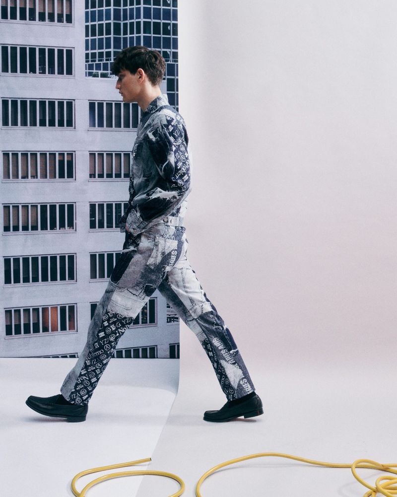 Young man in patterned outfit poses against a backdrop featuring a tall building and a plain white wall in a fashion shot.