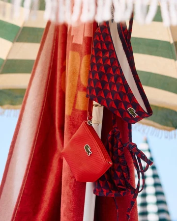 Lacoste branded swimsuit and pouch hanging under a striped beach umbrella. The red and blue swimsuit features a geometric triangle pattern.