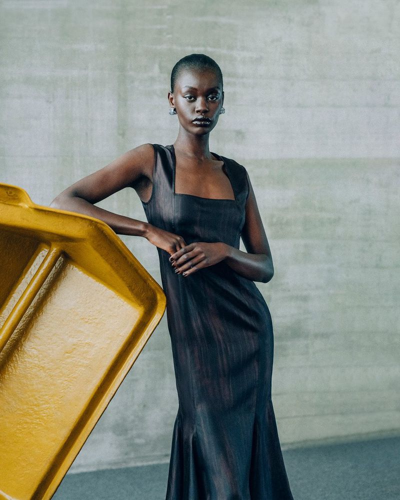 A slender Black woman in a sleeveless black dress leans on a yellow container against a concrete wall in a neutral studio shot.