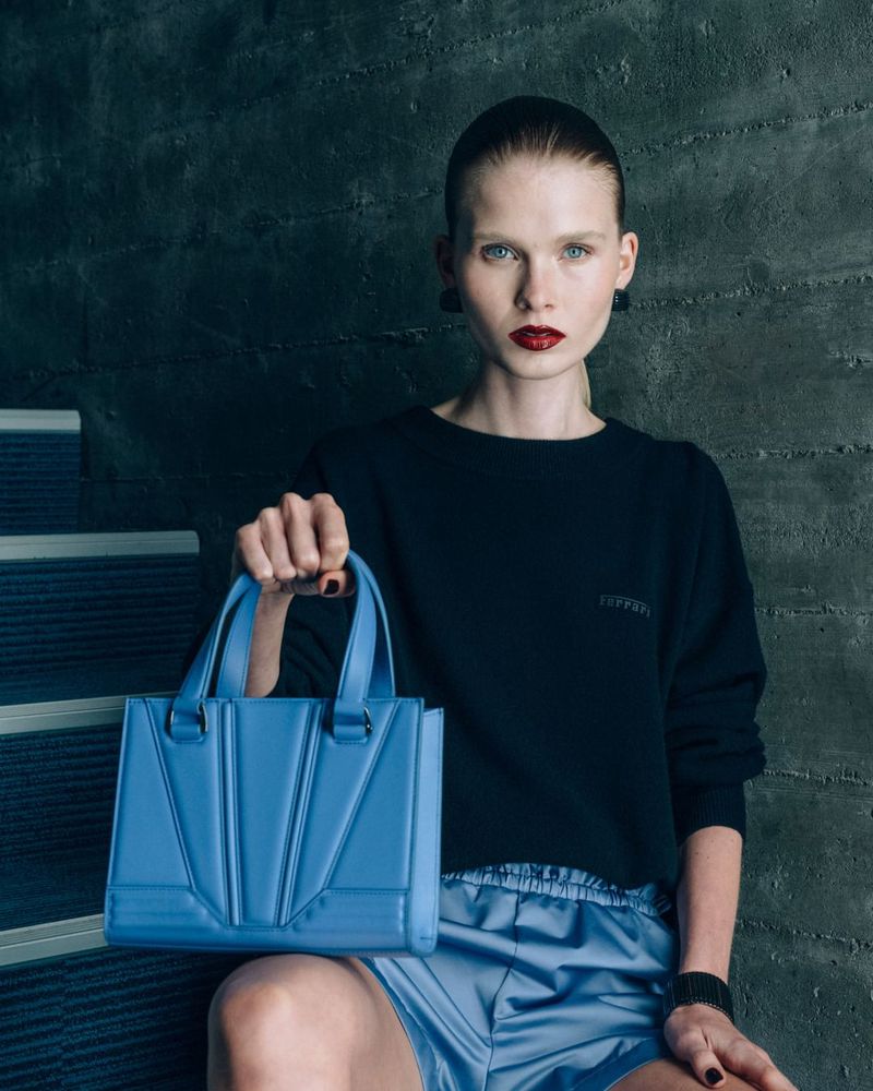 Model holding a blue leather handbag wearing a black sweater against a textured concrete wall for Ferrari.