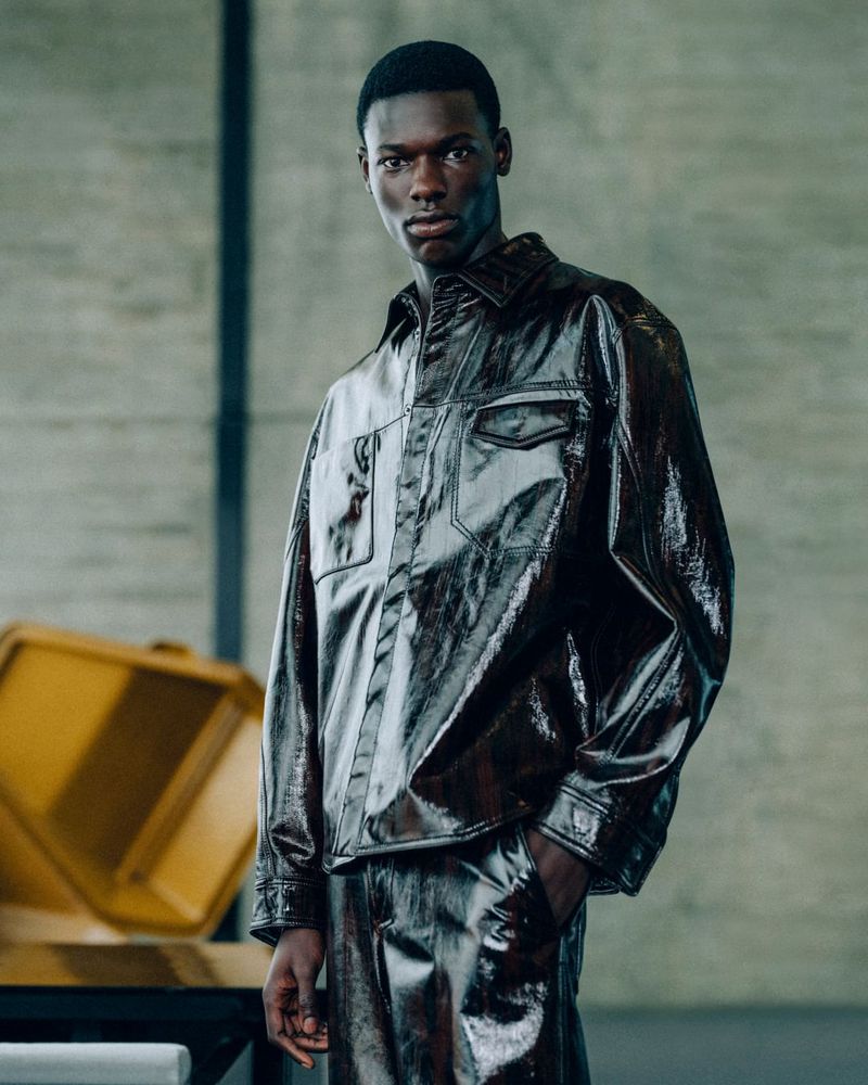 Stylish portrait of a Black man wearing a shiny, dark-brown two-piece outfit, posing in a minimalist concrete interior with soft natural lighting.