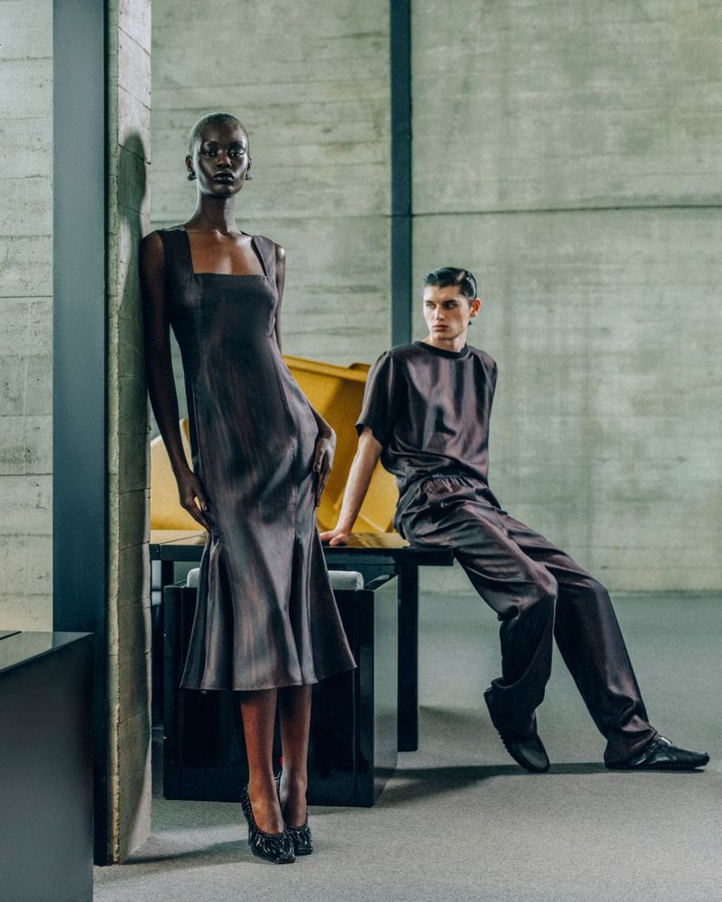 Two fashion models pose in a studio, one standing in a brown dress, the other sitting in a matching top and pants. Concrete background.