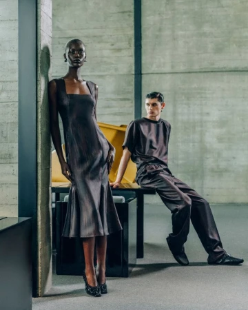 Two fashion models pose in a studio, one standing in a brown dress, the other sitting in a matching top and pants. Concrete background.