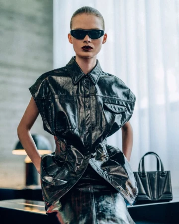 Stylish adult model in black sunglasses and avant-garde faux-leather outfit and handbag stands indoors against a gray wall and window.
