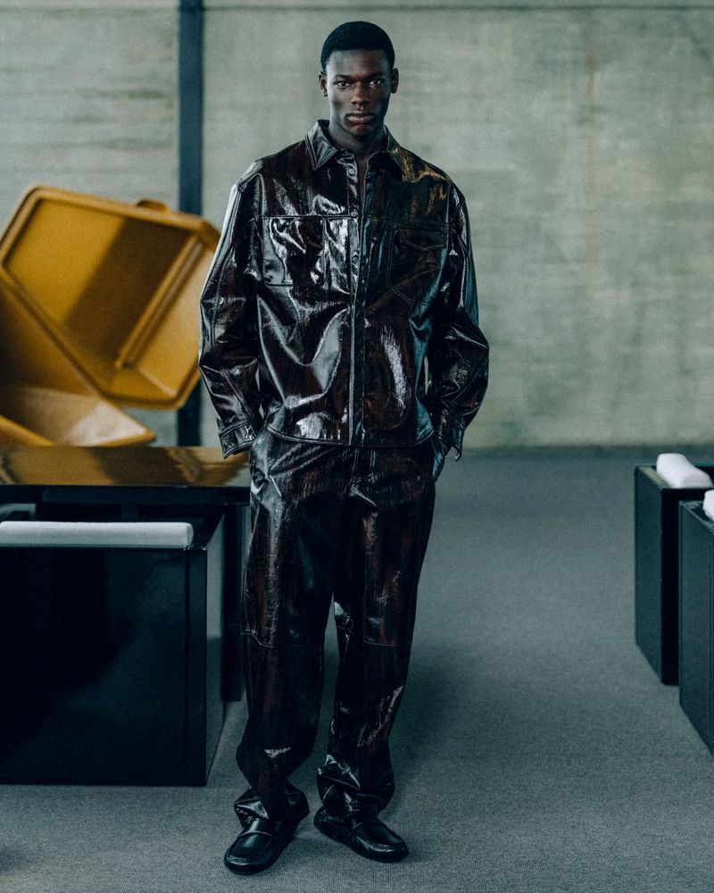 Fashion shot of a Black male model in a shiny brown jacket and matching pants, posing indoors with modern furniture and a concrete background.