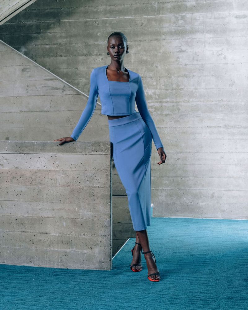 A Black woman in a blue, long-sleeved top and skirt poses next to a concrete staircase, creating a chic, modern fashion image.