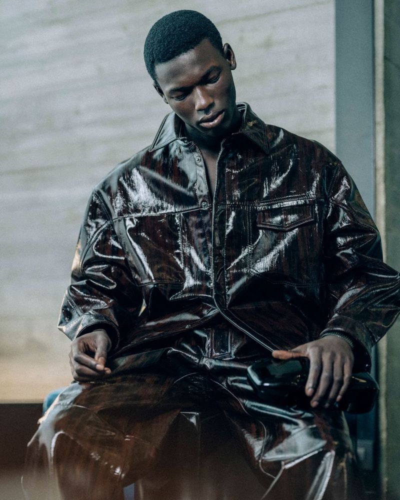 Fashion shot of a Black male model in a shiny brown shirt and skirt, posing indoors against a neutral-toned wall.