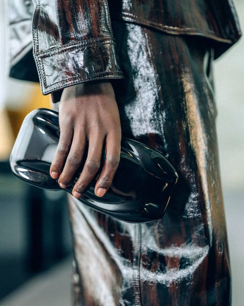 Close-up of a dark-skinned hand holding a glossy black clutch purse. The model wears a shiny, brown, vinyl jacket and skirt outfit.