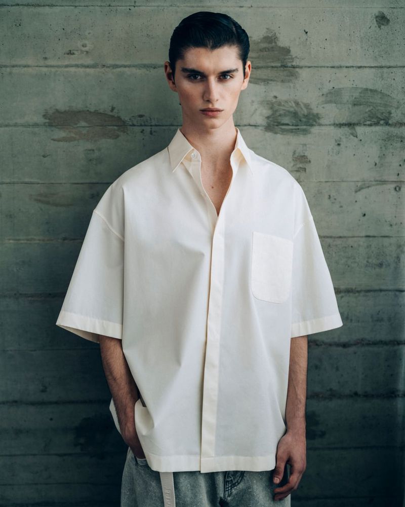 Portrait of a young male model in an oversized white shirt and grey jeans standing against a textured concrete wall in soft lighting.