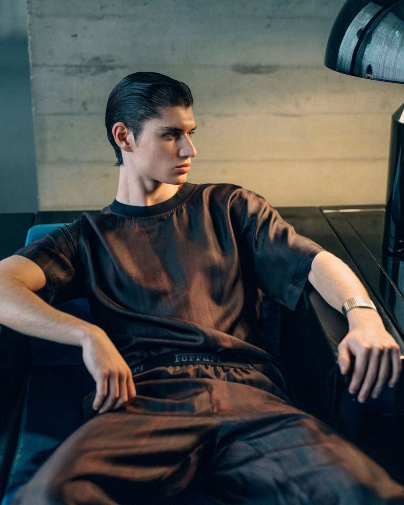 Portrait of a young adult male model wearing brown silky athletic suit, seated indoors with neutral grey background and professional lighting.